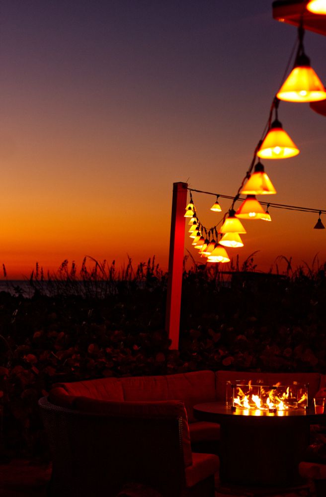 A string of yellow tinted, conical lights are hung over a fire pit at sunset. The sky is a beautiful gradient of red, orange, yellow, and violet. Silhouettes of plants are sticking up from the ground behind the fire pit.