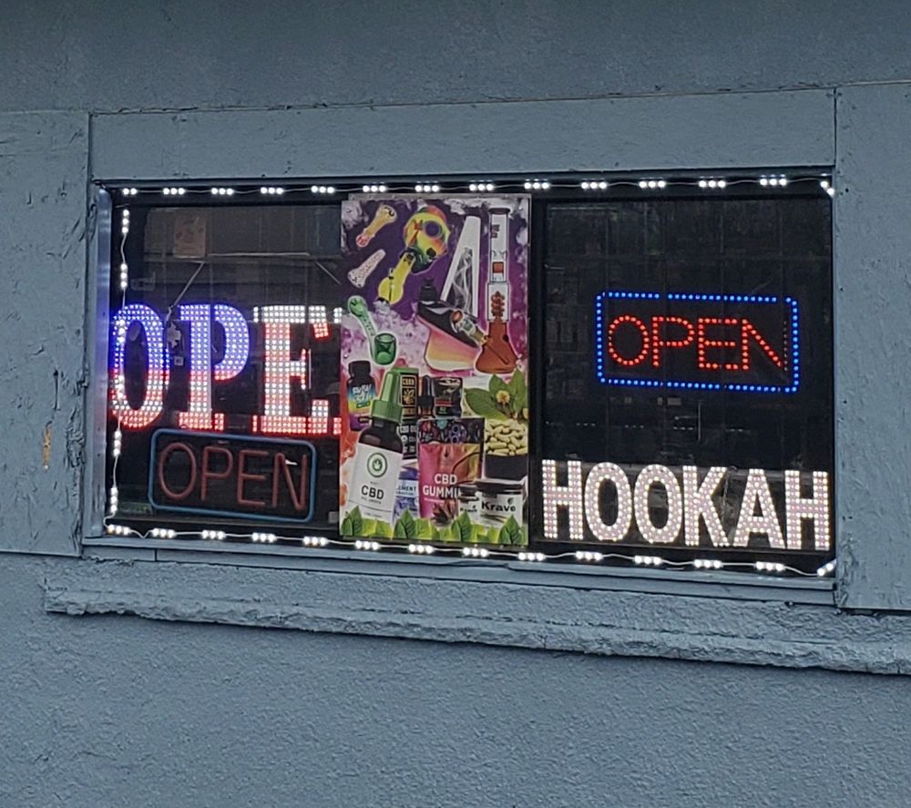 A hookah shop window that has a poster of smoking apparatuses in the middle. To the left are two open signs: one that isn't lit and one that has LED lights to make it display the American flag, which is partially blocked so it looks like it just says "OPE." To the right is yet another open sign, which is on, and a sign that says "HOOKAH."
