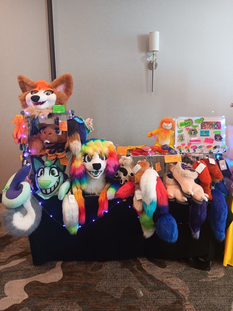 A booth nearly overflowing with fursuit parts, partials, and various furry merchandise. We are all set up with YCH market!!