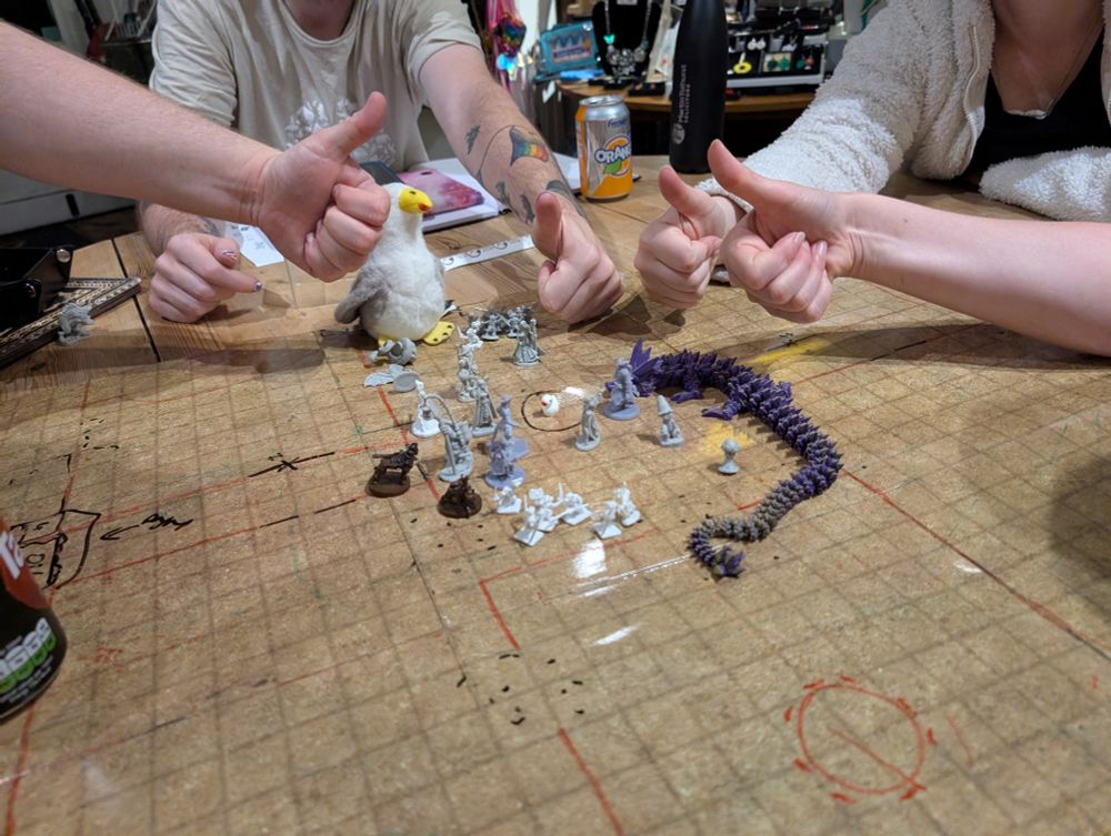 A dry wipe battle map with loads of minis, a 3 d printed dragon and a small plush seagull. Around the map, four people's hands giving a thumbs up