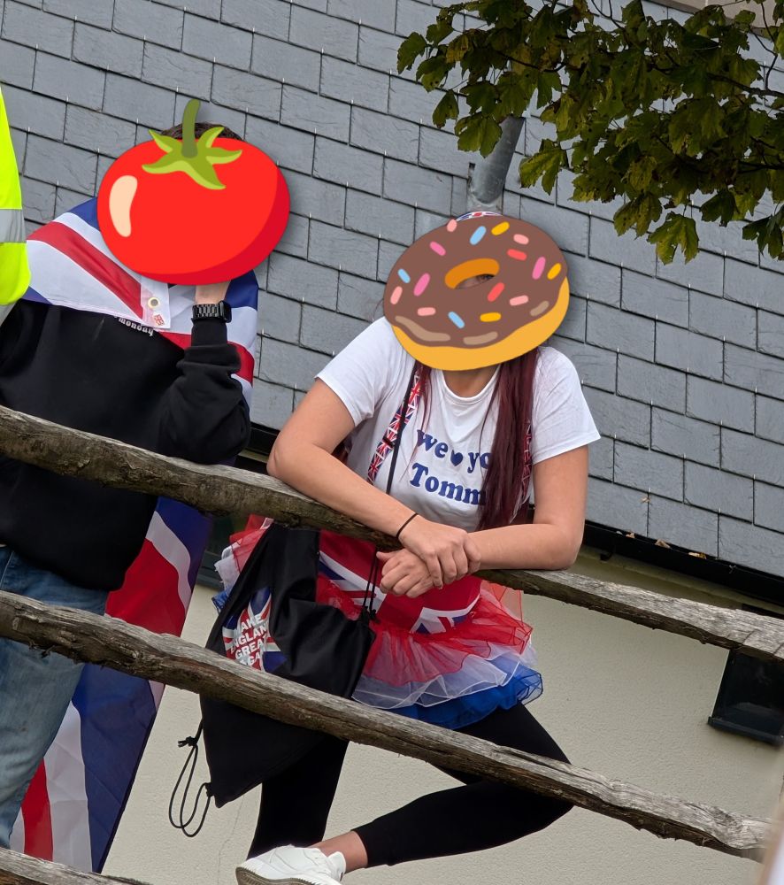 A couple of anti immigration protesters with their faces obscured by emoji. Behind a tomato is a young man with a union jack cape. Behind a doughnut is a young woman wearing a union jack tutu, a tote bag with "make England great again" on it and a t-shirt with "we ❤️ you Tommy" on it 
