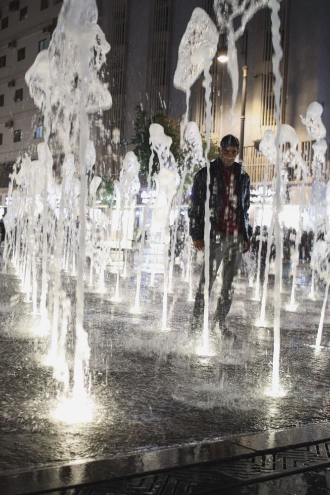 Plumes of uplit water surround a man as he poses in the middle of a street level water feature. 