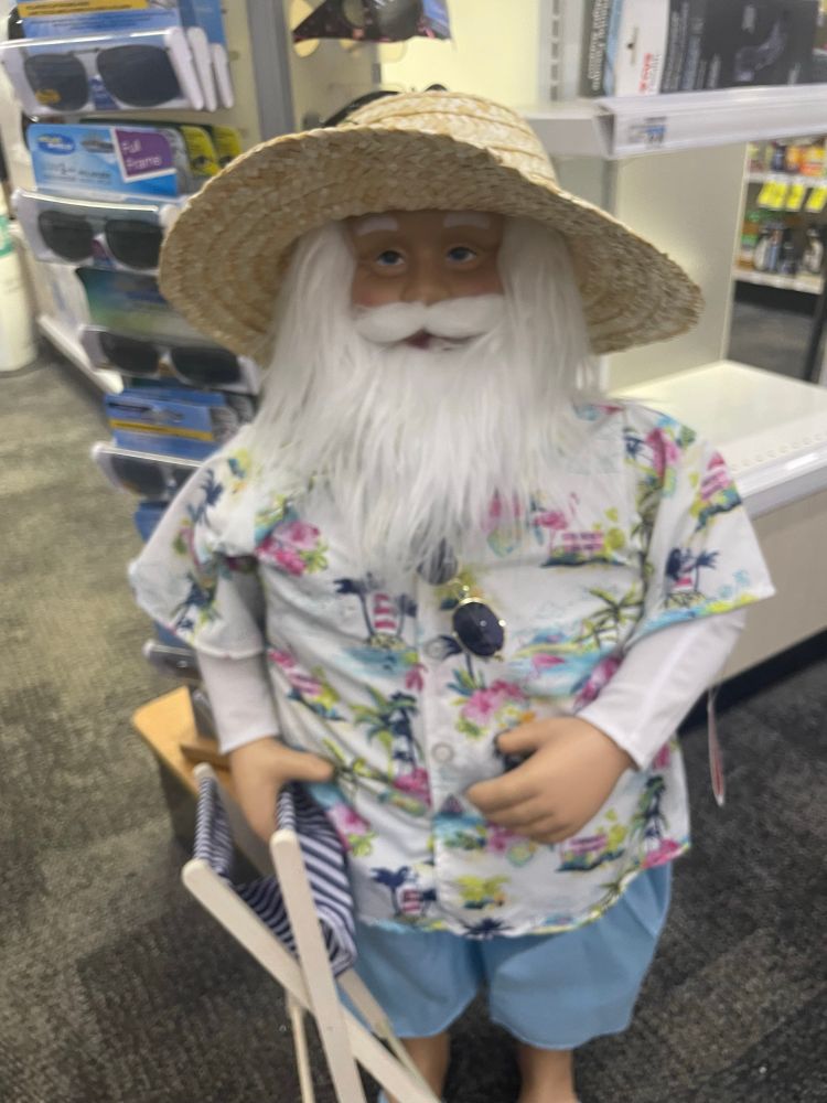 Santa in a straw hat holding a beach chair. He has sunglasses hanging off his Hawaiian shirt, which has palm trees and lighthouses on it 