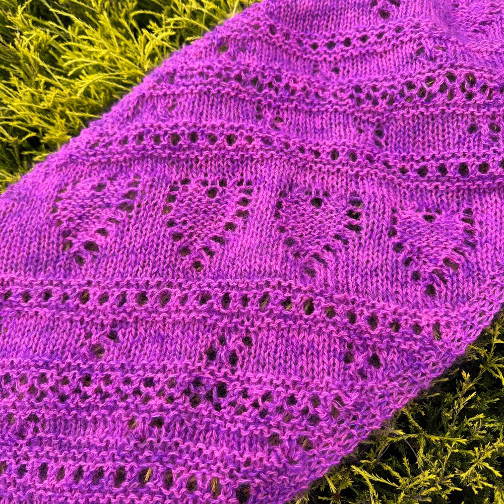 A bright handknit pink scarf laying over some green plants showing a portion of it, the portion displays heart motifs and lacy sections.