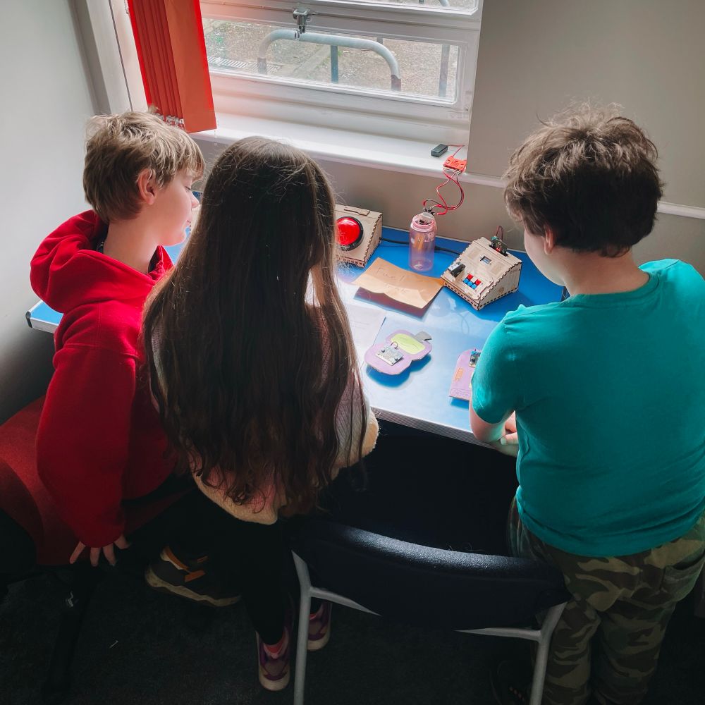 Three children taking part in a microbit based escape room activity, with cardboard robots and wooden computer consoles on a table in front of them. 