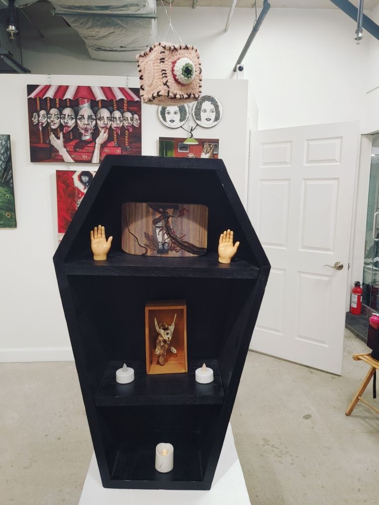 Coffin Shelf/convertible cat bed by Michael Giel. Two small art pieces on shelf including bones and dessicated snake by Shona Chornenki. Crocheted eyeball cube by CuteCreepyCrochet. Conjoined Twins by Vashty Hawkins. Creepy Carousel with faces and tongues by Anika Becker.