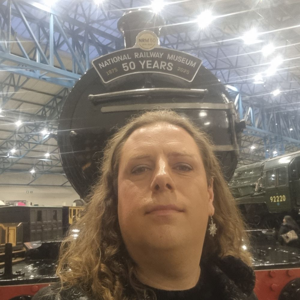 Wavy brown haired white trans woman in front of a steam locomotive with a sign saying national railway museum 50 years.