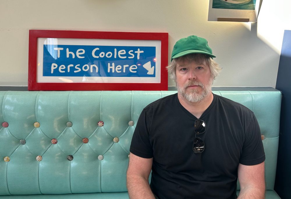 A 40-something man in a green hat and black shirt sits in front of a sign that reads The Coolest Person Here