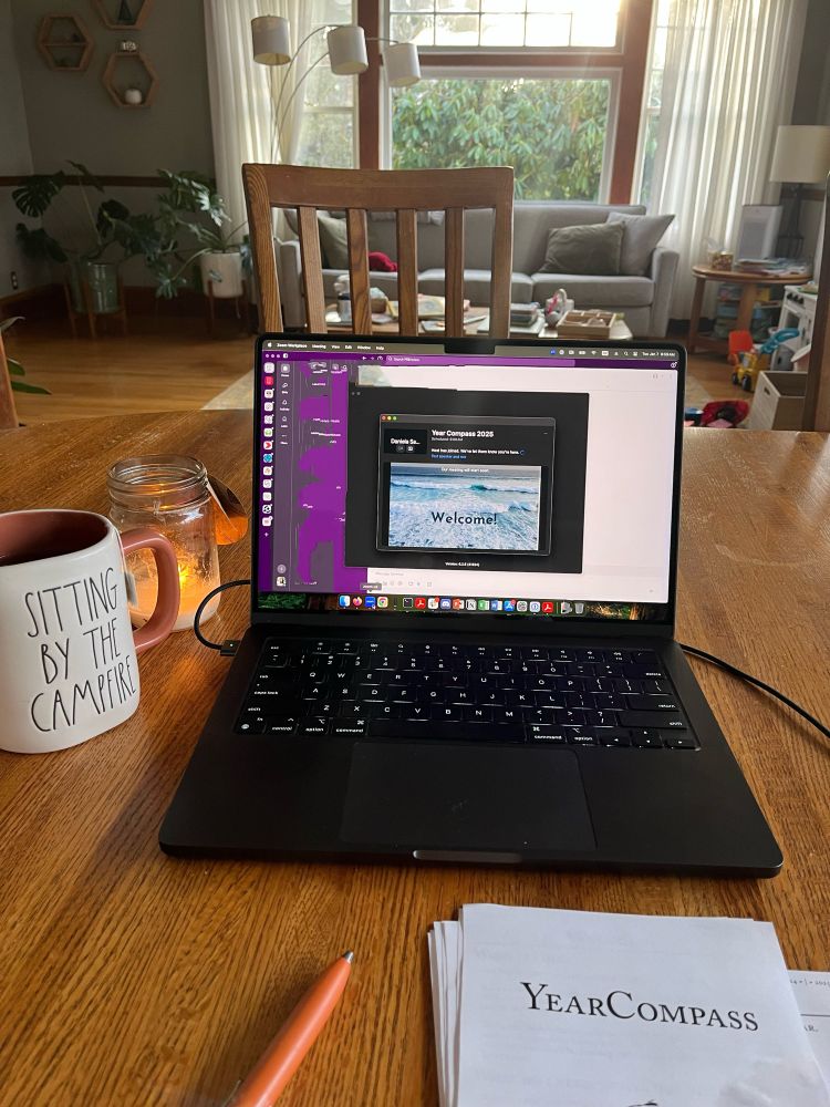 Picture of a black computer on a wooden table with the waiting room zoom window open in the center-written in it is the word Welcome! on top of a picture of the ocean waves. Next to the computer is a tea mug with “Sitting by the campfire” written on it and below the computer are an orange pen and the printed copy of my YearCompass booklet.