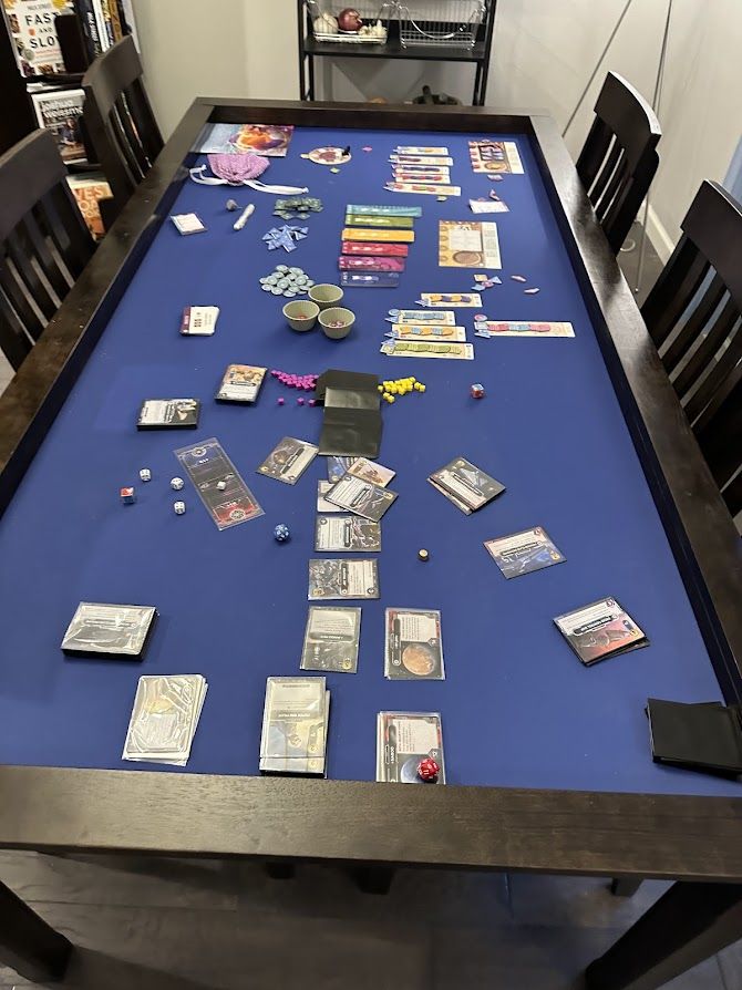 Vertical shot of a long board game table, with two games. Foreground is Star Wars The Deck Building Game. Background is Knitting Circle. It's a dark wook table with chairs on either side. The table top is inset for gaming with a blue top.