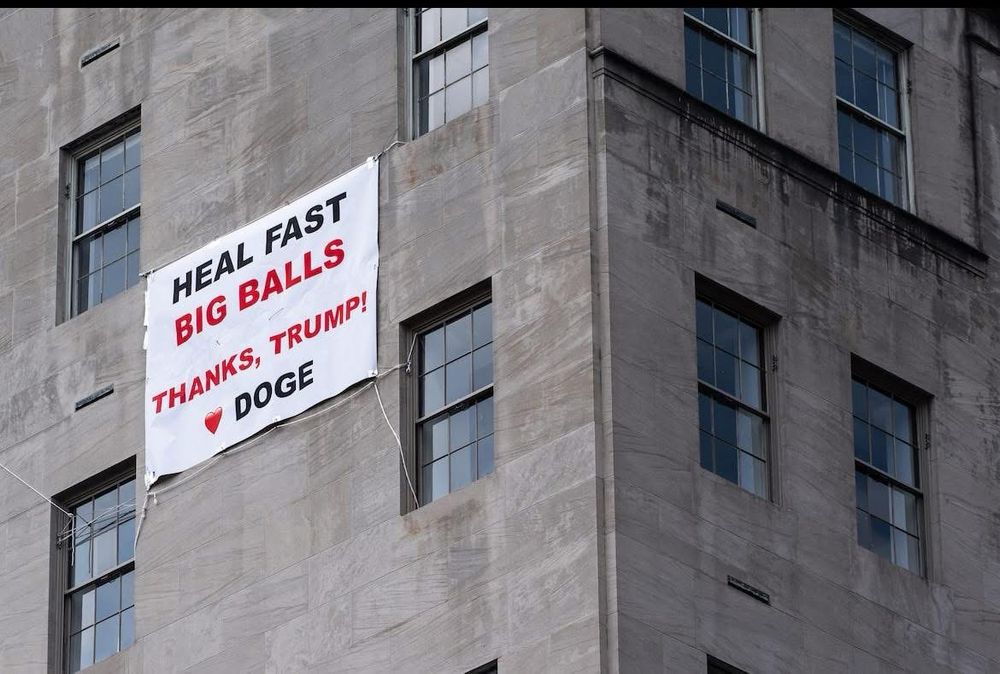 A banner hanging from some windows. It says, incredibly: HEAL FAST BIG BALLS 

THANKS, TRUMP! 

❤️ DOGE 
