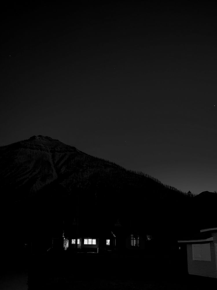 A black and white photo of a mountain at twilight. Three stars dot the sky over the mountain. A small cabin sits in front of the mountain, its windows aglow with light piercing the darkness.  