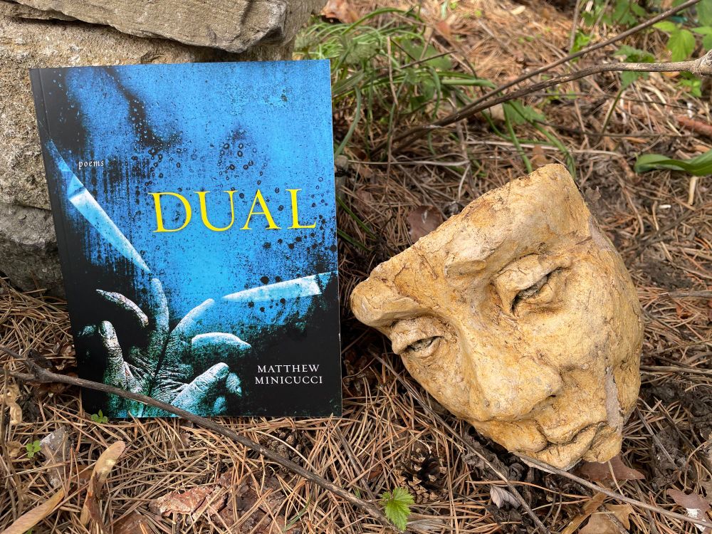 Matthew Minicucci's poetry collection, DUAL (blue cover with two hands and knives), rests beside a dramatic mask.