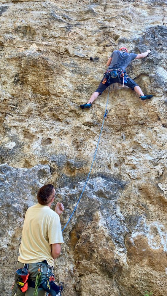 Two figures in front of a tan and grey limestone rock face. They are rock climbers. In the lower left a person in a yellow shirt belays the climber in the upper right. Who is obviously sweaty, situated with legs wide apart on the footholds, and reach further up and to the right. They both have an assortment of equipment attached to their harnesses.