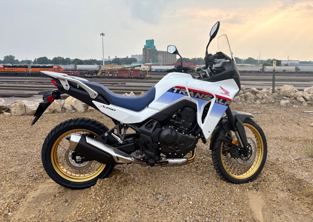 A white, blue, and red Honda Transalp motorcycle is parked on some gravel in front of some train tracks. The sky is hazy with crepuscular rays.