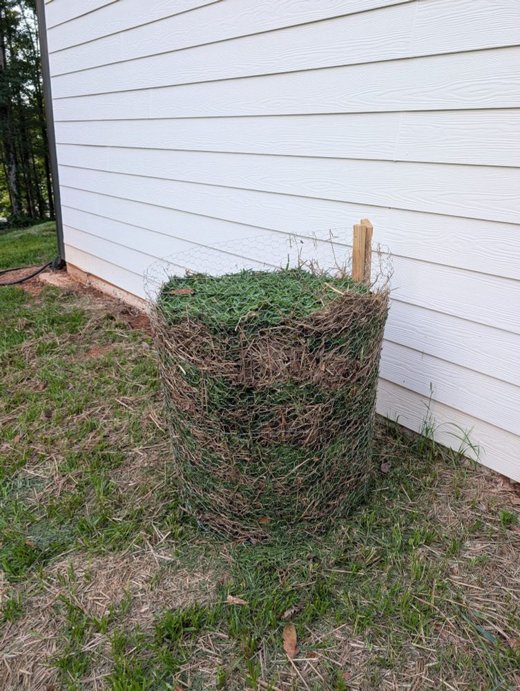 Lasagna like layered compost with grass and hay layers in a chicken wire circular compost. About 3ft tall and 2.5ft diameter. White house behind with shoddy grass around it.