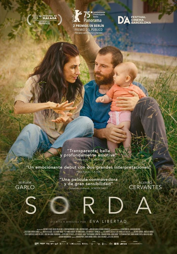 Spanish poster of the film “Sorda”. Young parents and their baby are sitting in the grass. The father is leaning against a tree and holding up the baby. Both are looking at the mother, who is holding eye contact with her baby while signing.