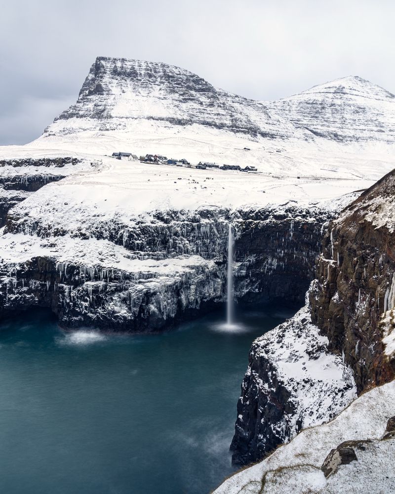 The scene from the Faroe Islands is dominated by a massive snow covered mountain rising from the cold dark blue Atlantic Ocean with with a gorgeous waterfall in the center of the scene streaming into the ocean.