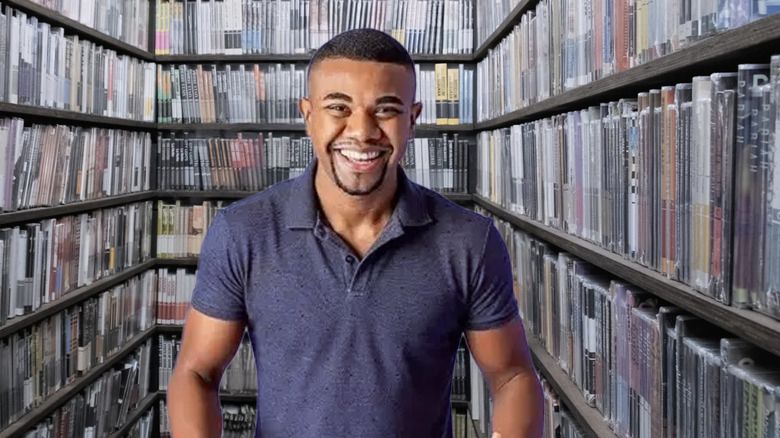 davi brito com uma camisa polo roxa sorrindo dentro do closet da criterion rodeado de dvds e blurays de filmes 