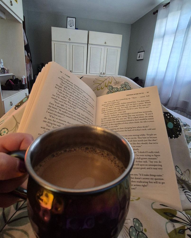 A light green painted bedroom with white wardrobes in the background with a curtain covered window to the right. The book Somewhere Beyond the Sea by TJ Klune is open and resting on knees covered in a flowery duvet with a cup of coffee held in front of it.