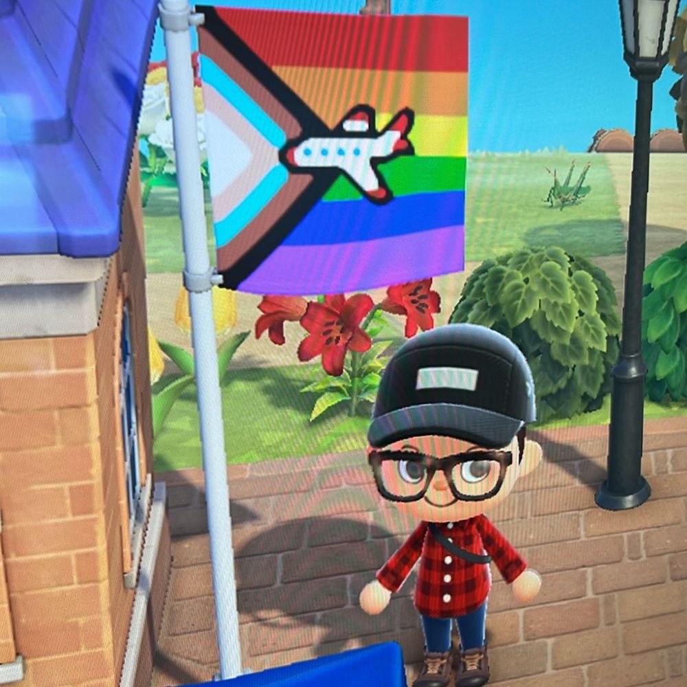 My Animal Crossing avatar standing next to the town square flag pole. I’m wearing very queer flannels with a black cap and brown boots. The flag on the pole is the queer inclusion flag with a big plane in the centre. 
