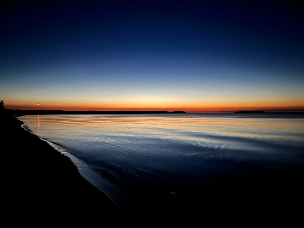 Dusk over a calm, shallow inlet of Lake Superior. A crescent moon is visible on the left horizon.