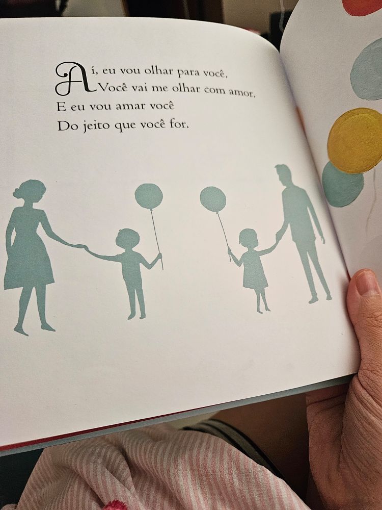 Livro infantil aberto, silhueta de dois adultos e duas crianças segurando balão. No livro o texto "Aí eu vou olhar para você. Você vai me olhar com amor.  E eu vou amar você do jeito que você for." Trecho final do livro As Maravilhas que Você Vai Ser, de Emily W Martin.