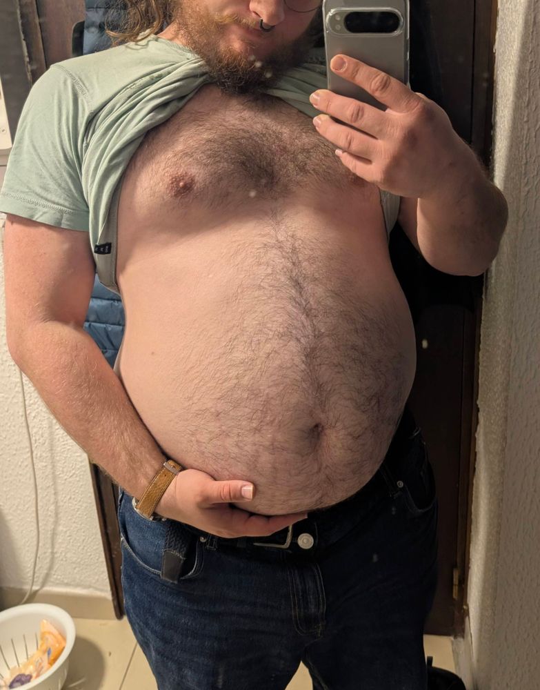 A mirror selfie of me, shirt pulled up and wearing jeans, showing off my big belly while holding its underside in my hand.