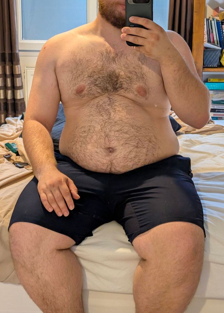 A mirror selfie of Orme sitting on his bed, shirtless a'd wearing form-fitting gym shorts.