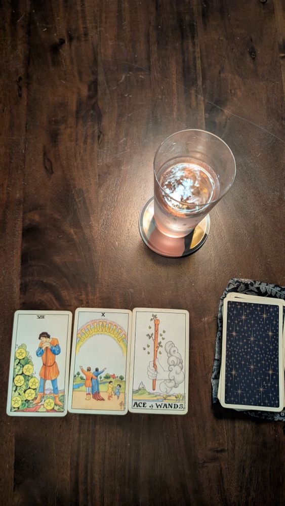 A glass of sparkling water sits on a coaster near a pile of unturned, starry, tarot cards. Three have been pulled, left to right all upright: 7 of cups, 10 of cups and ace of wands.