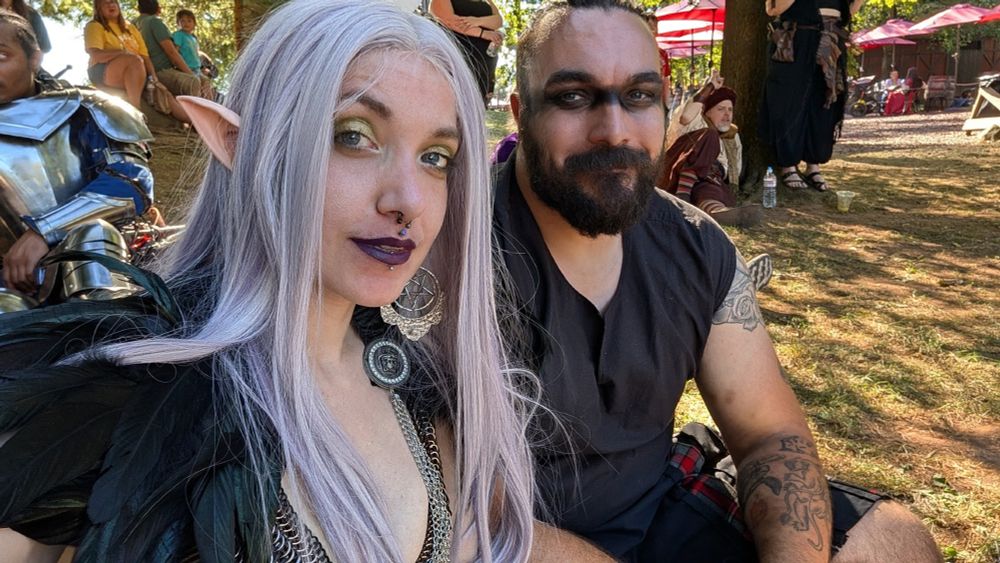 The image shows two people sitting outdoors on a sunny day at a Renaissance Fair. The person on the left has long, pale purple hair and is wearing green eye shadow, a dark purple lip color and costume with elf ears and a black feathered mini cape. The person on the right has a beard, dark eye makeup, and tattoos on their arm, wearing a black sleeveless top. In the background, there are other people in fantasy & period costumes, both seated & standing under trees and near red patio umbrellas. The ground is covered with grass and dirt, and sunlight filters through the trees, creating dappled lighting.