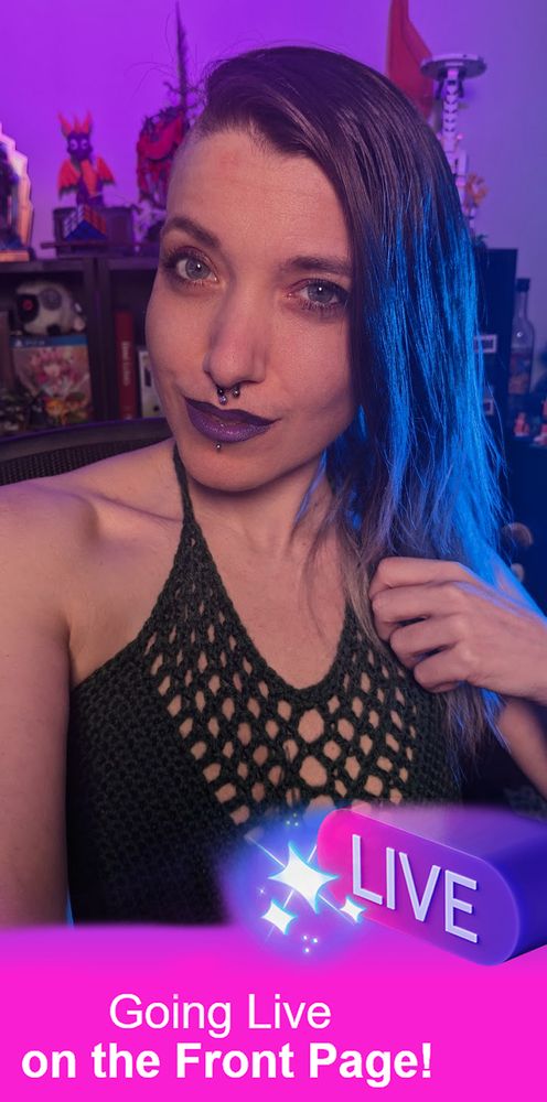 The image features Amaris, a person with long, dark brown to grey ombre hair wearing a dark green crochet top, posing indoors. The room is softly lit with purple lighting. In the background, a shelving contains various items, including books and decorative figures. Amaris is wearing dark purple lipstick and has facial piercings. They have a confident expression with a slight smile. On the lower part of the image, there is a vibrant pink banner with a glowing "LIVE" button and white text overlay stating "Going Live on the Front Page!"
