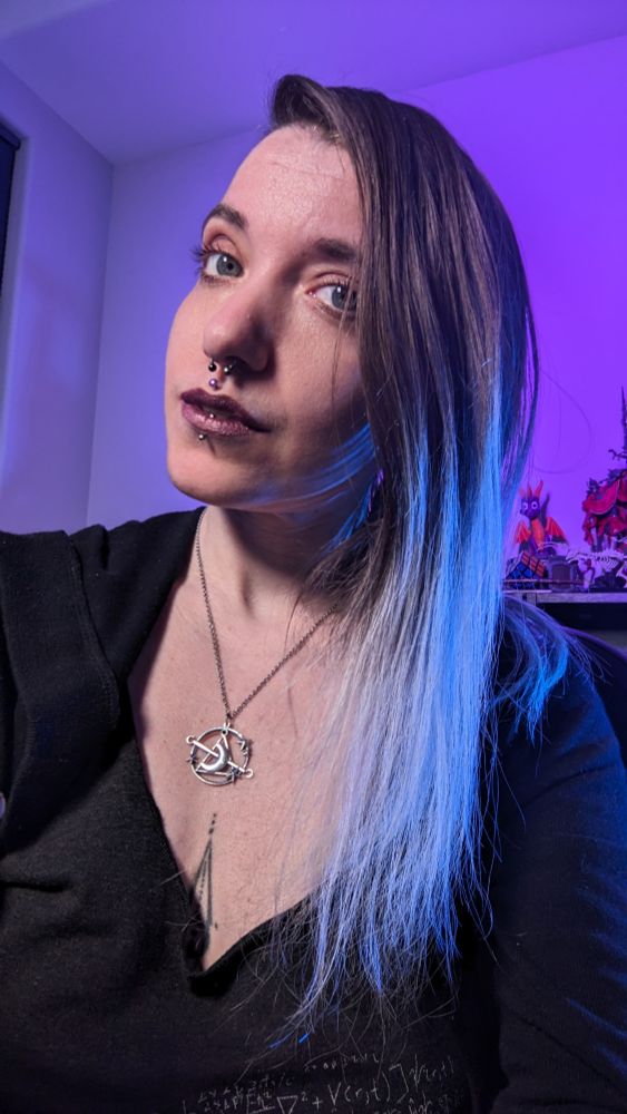The image features Amaris, a person with long, straight hair that transitions from dark at the roots to light towards the ends, highlighted by purple and blue lighting. They have a septum, philtrum and vertical labret piercing and are wearing dark metallic red lipstick. Amaris is dressed in a dark grey tank with a black sweater over top and wears a silver necklace with a circular pendant representative of Nyx. In the background, there are colorful figurines and collectibles displayed on a shelf. The overall lighting gives a soft purple and blue hue to the scene.