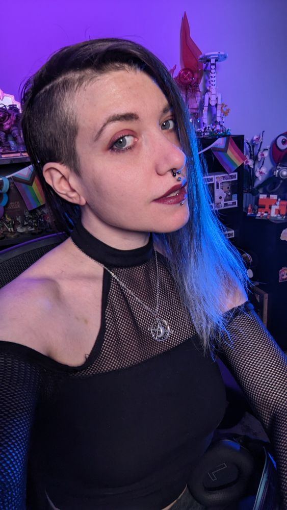 The image shows Amaris, a person with a partially shaved hairstyle, featuring long ombre brown to grey hair on one side. They have light skin and are wearing a mesh black top with a high collar and off-the-shoulder sleeves. A silver pendant necklace with a symbol for Nyx adorns their neck. Amaris has facial piercings, including a septum, philtrum & vertical labret piercing. In the background, there's a shelf with various items, including a progress pride flag bunting and several collectible figurines. The lighting in the image is purple with a blue side lighting.