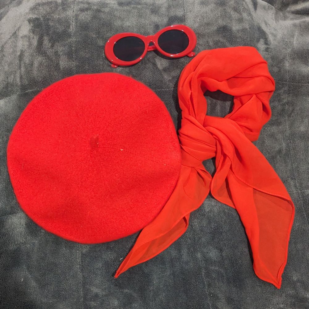 The image features three red fashion accessories laid out on a dark grey, textured fabric background. On the left is a round, vibrant red beret made of soft wool. To the right is a translucent red scarf, elegantly draped and knotted, displaying a fluid, lightweight texture. Above these items are a pair of rounded red sunglasses with dark lenses.