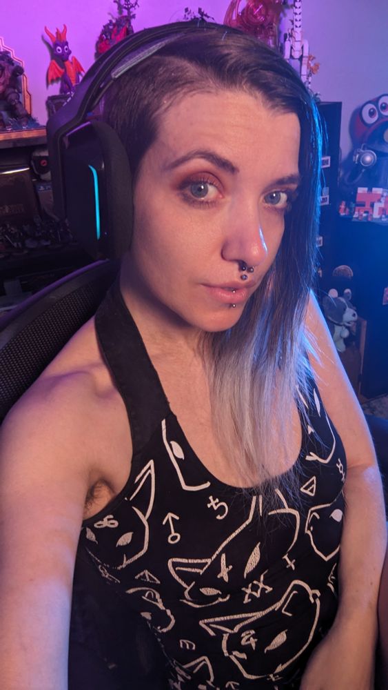 The image shows Amaris, a person with long hair, partially ombre dyed brown to silver, sitting indoors and wearing large over-ear headphones with a subtle illumination visible on the ear cup. Amaris has facial piercings including a septum, philtrum and vertical labret. They are wearing a black top with a white design of cat head and occult symbols. The background is a room with colorful lighting and shelving holding video game collectibles.