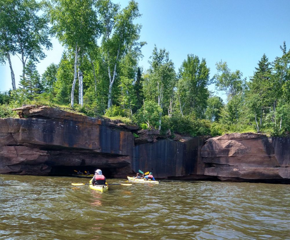 two kayaks on a lake in front of red rock formations, with one kayaking emerging from a sea cave in the rock