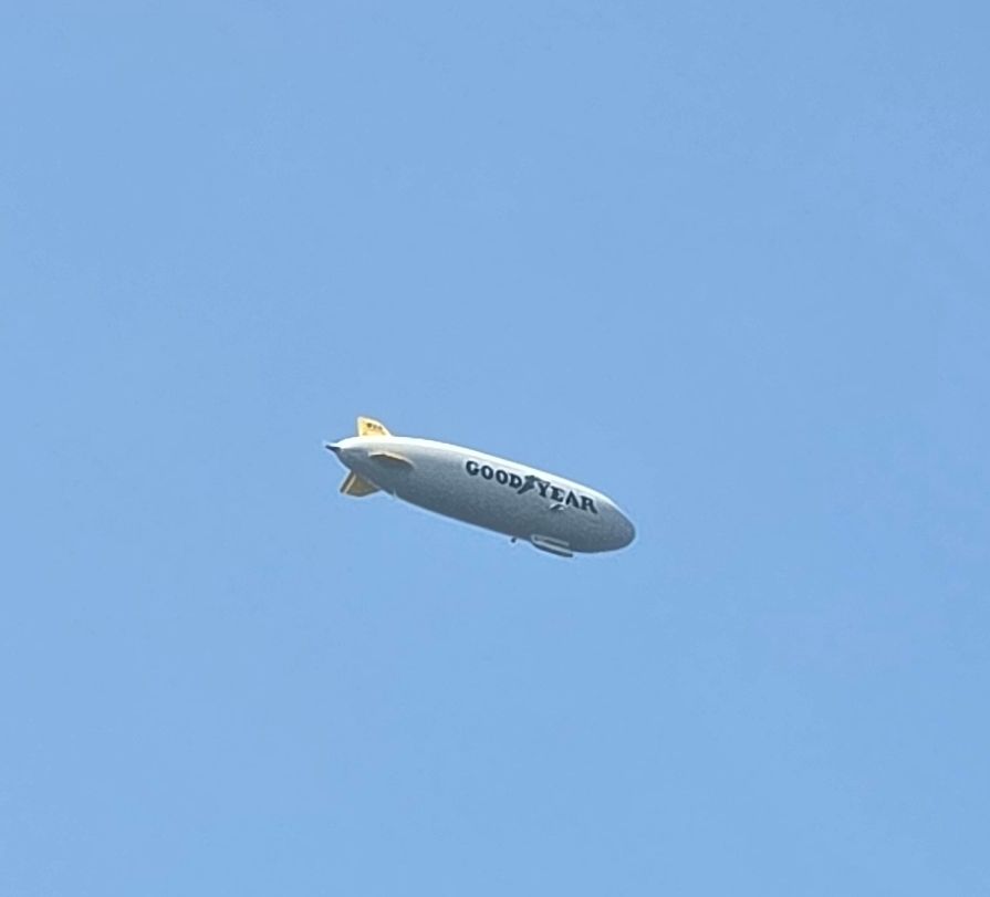The Goodyear Blimp flies in a blue sky above trees