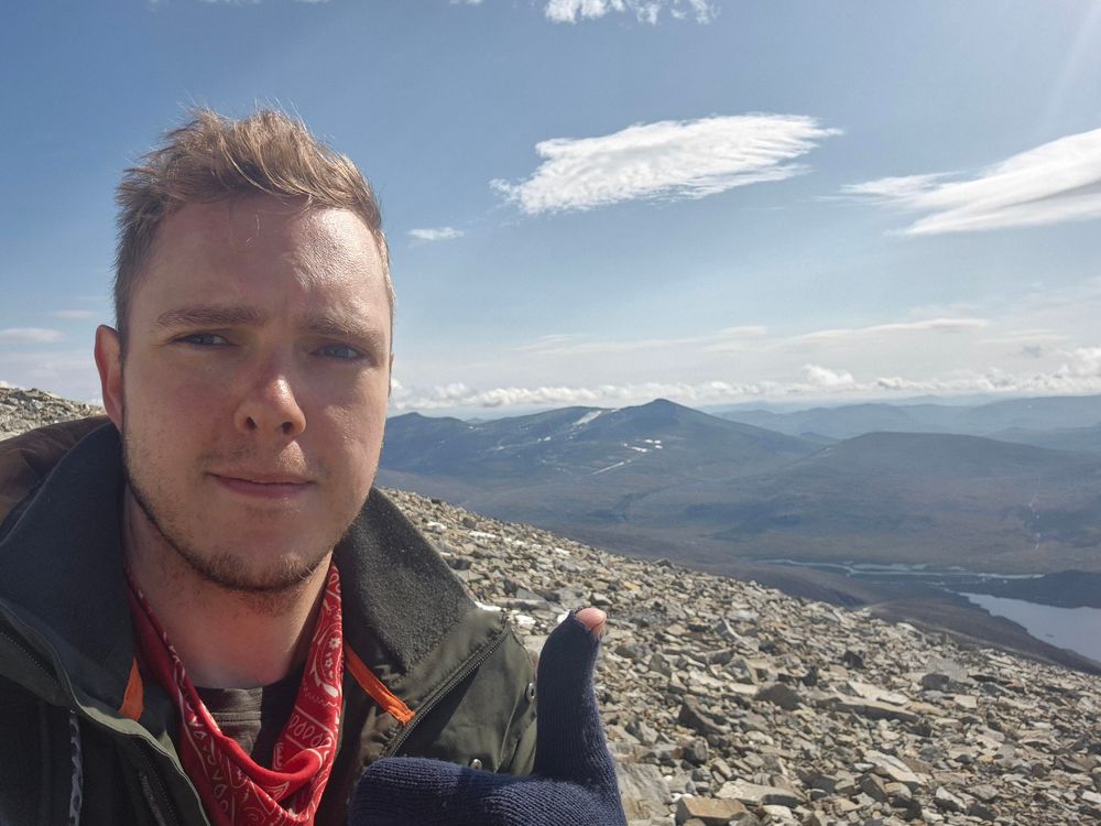 a selfie with me doing a thumbs up on Glittertind with the background towards Glitterheim and various mountains in the background 