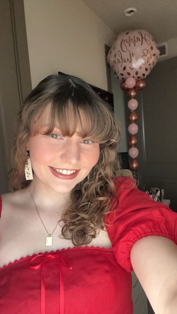 A selfie of me. I’m wearing a red dress with my gold rectangular necklace hair pulled to the side and one earring visible. There are pink balloons over my left shoulder saying Happy Birthday 