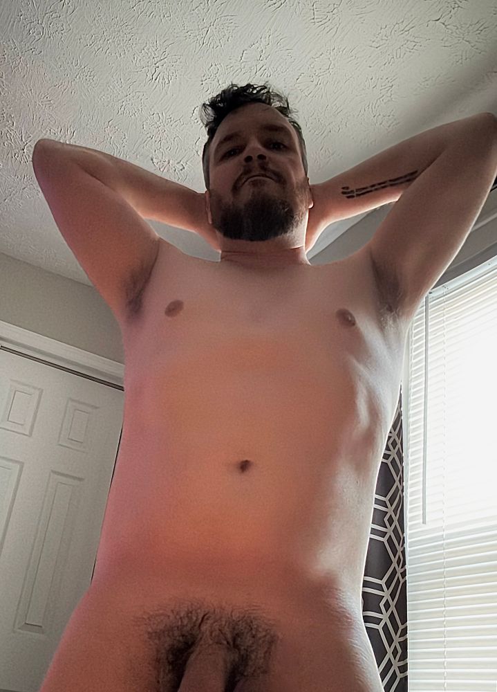 Beefy bearded cis white dude poses naked with his arms up, exposing his hairy pits; his hairy bush and root of his thick cock are visible