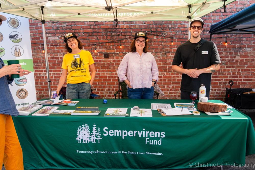 Three people standing behind a booth with the "Sempervirens Fund" banner, smiling at an outdoor event. Various informational materials are displayed on the table.