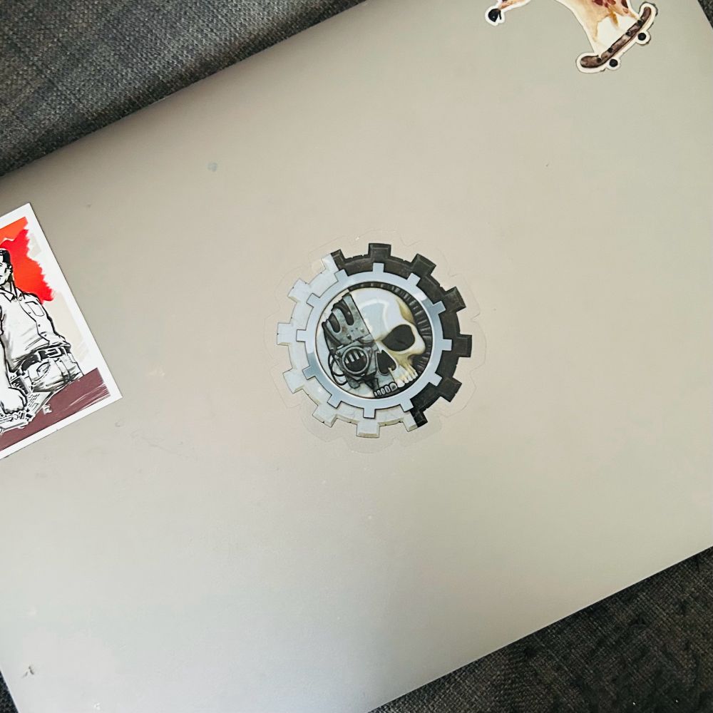 A laptop with three stickers: a black and white illustration of a person with a red background, a skull inside a gear, and a giraffe on a skateboard.