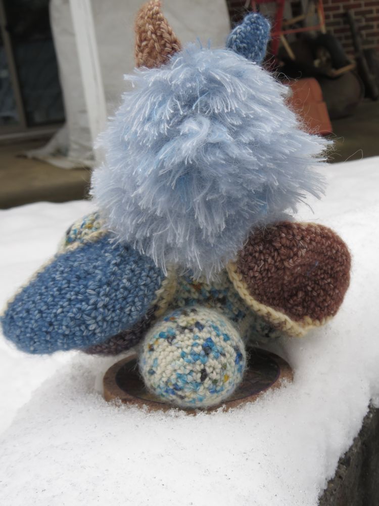 A colourful crocheted moth sitting outside in the snow, showing off the wings
Main colours are off white, blue and brown.