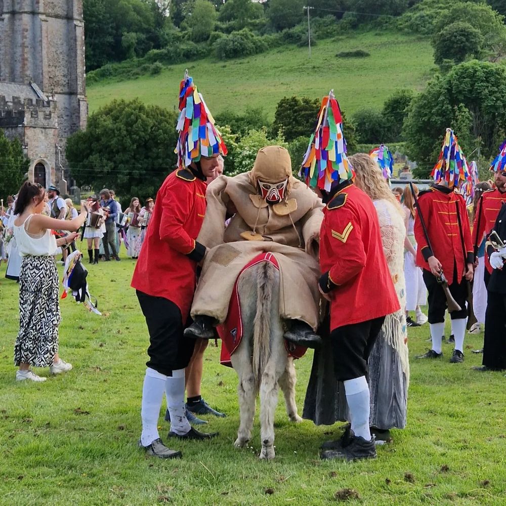 A character in a mask and sack costume is places on a donkey by two men in military style red coats with colourful pointy hats. There is a green hill and a church tower in the background and many people standing around