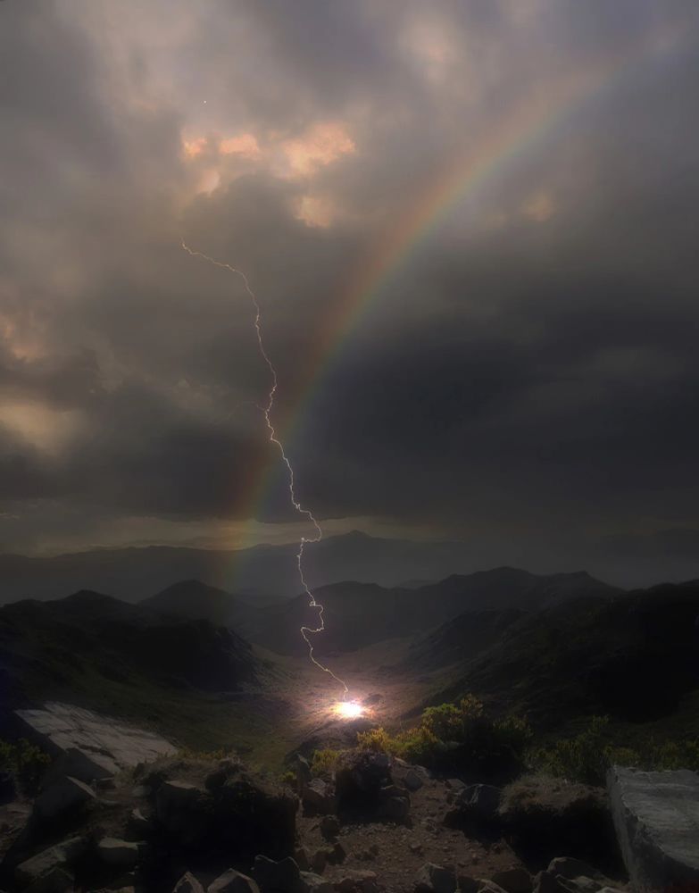 An impressive photograph of a mountainous landscape with dark clouds overhead. In the centre, a slim, jagged bolt of lightning breaks through, casting a patch of blinding white light onto the earth's surface. The lightning bolt is crossed over by a hazy rainbow. 
