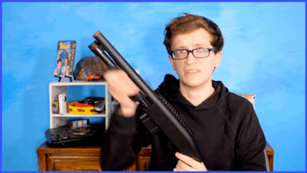 a man wearing glasses is holding a shotgun in front of a blue wall