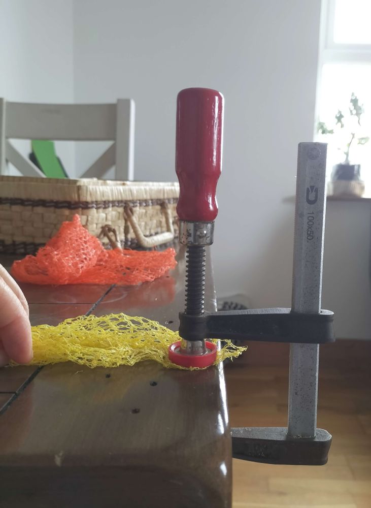 A clamp is holding a net securely in place. The net is a bright yellow colour. An orange coloured scrap if net is also shown. On the table is a basket full of hand - sewing threads and needles. 