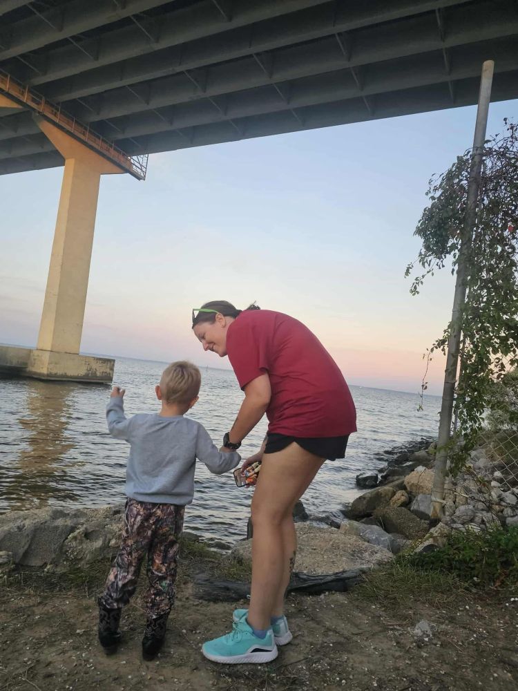 Woman and small boy stand at a rocky edge of the Gulf of Mexico, under the end of a bridge. The boy is facing the water and gesturing to it with one hand; the other hand is held by the woman, who is hunched over and talking to him.