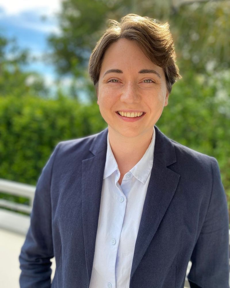 Ariel Marcy stands outdoors, smiling warmly. She has short brown hair and wears a dark blue blazer with a light collared shirt.